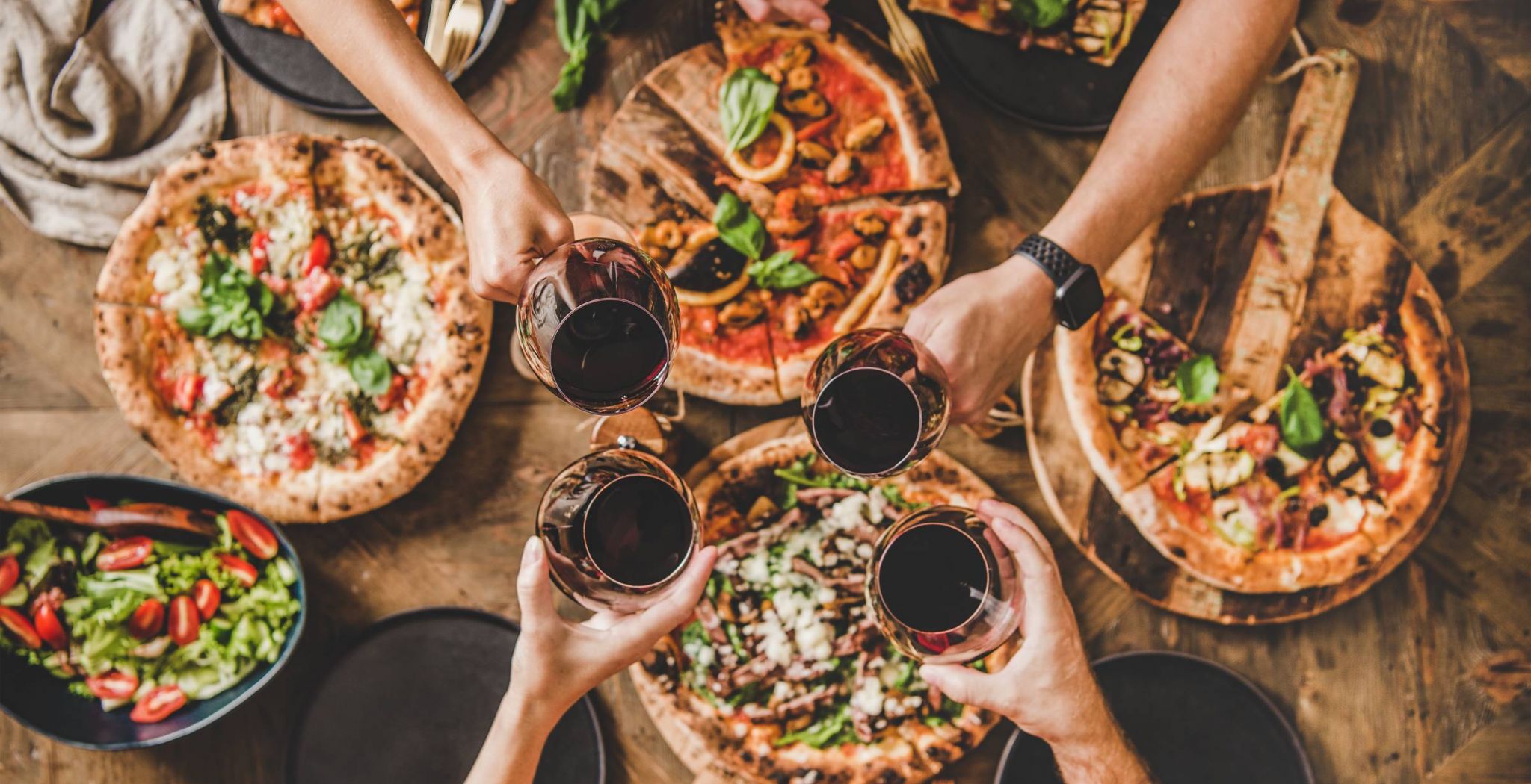 Overhead view of friends clinking wine glass over a table of pizzas.