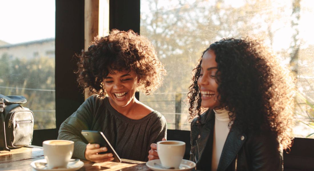 Two women laughing looking at a mobile phone inside a coffee shop. Friends sitting with coffee cups on the table having fun.